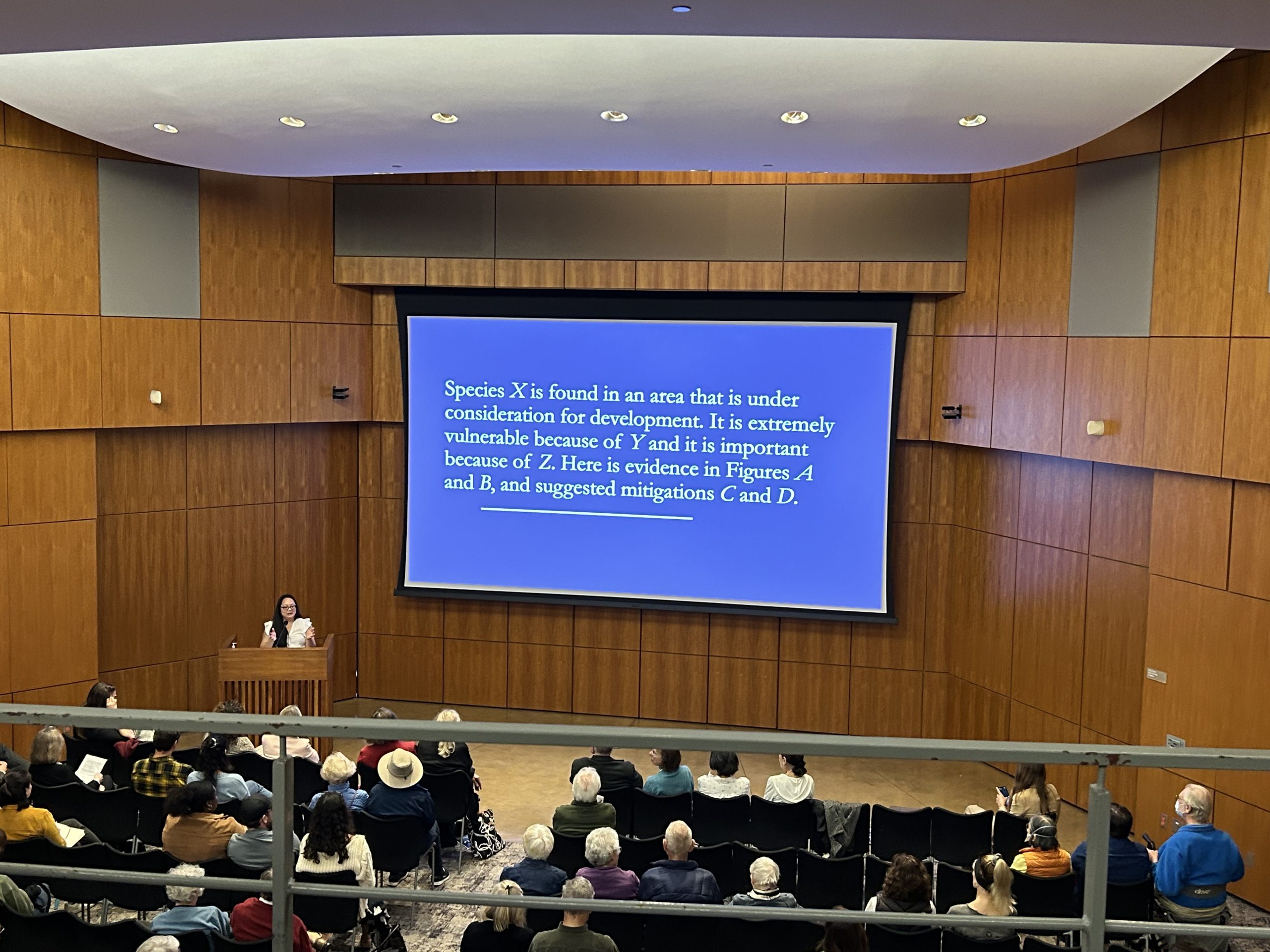Lecture hall with projector featuring a blue screen and text. A crowd sits in the foreground of the image, and a woman (Dr. Jen Le) located on the lower left of the image is presenting. 