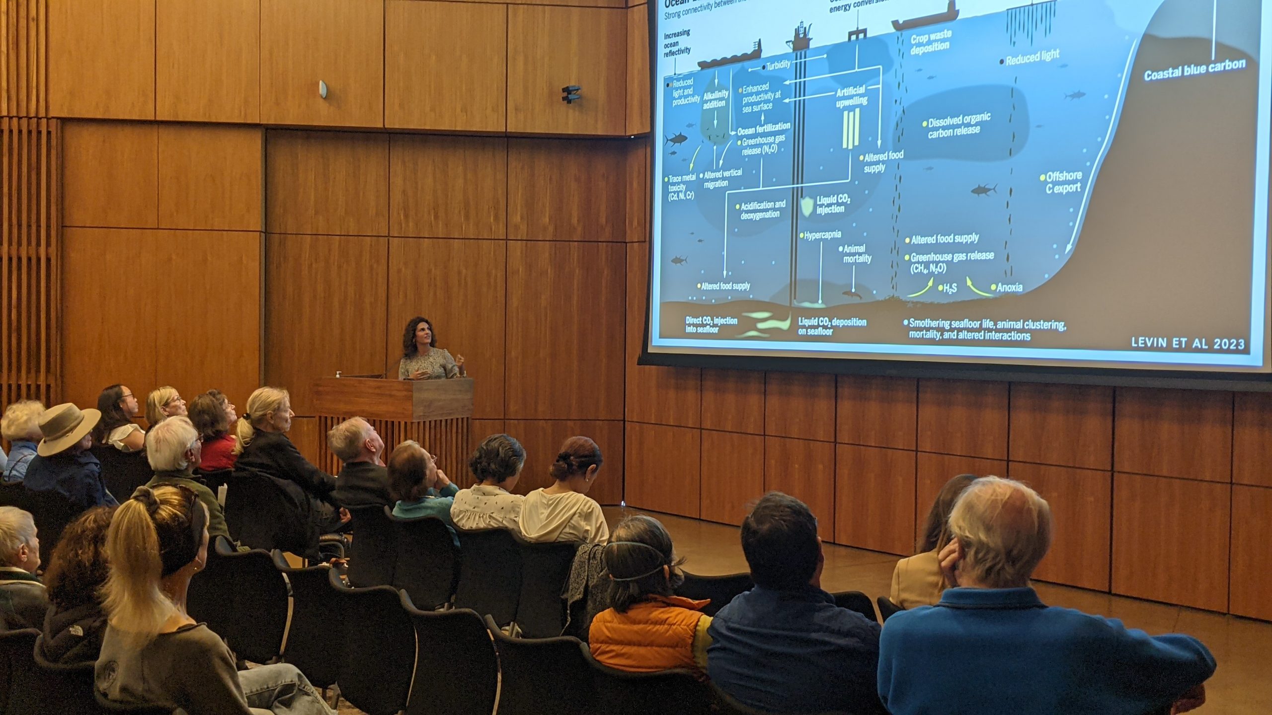 In front of crowd in the foreground, a woman (Dr. Diva Amon) presents in a lecture hall. She stands to the left of a projector screen featuring a diagram of the deep ocean.